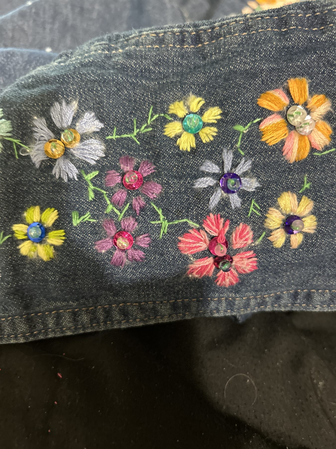 Denim cuff covered in small embroidered flowers in yellow, coral, lilac and orange, each with a sequin or rhinestone center and green stems.