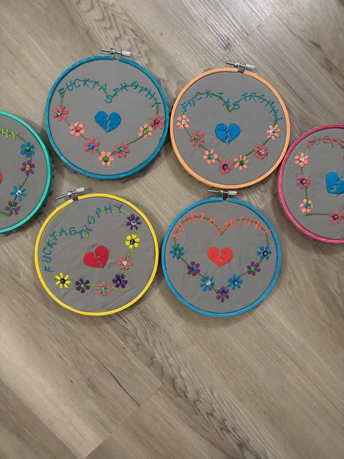 Six embroidery hoops laid out on a pale wood floor, each stitched with the word Fucktastrophy curving into the shape of a heart around a broken heart at the center. The hoops are painted teal, orange, yellow, blue, pink and green, and each has its own colorway of flowers with sparkling centers.