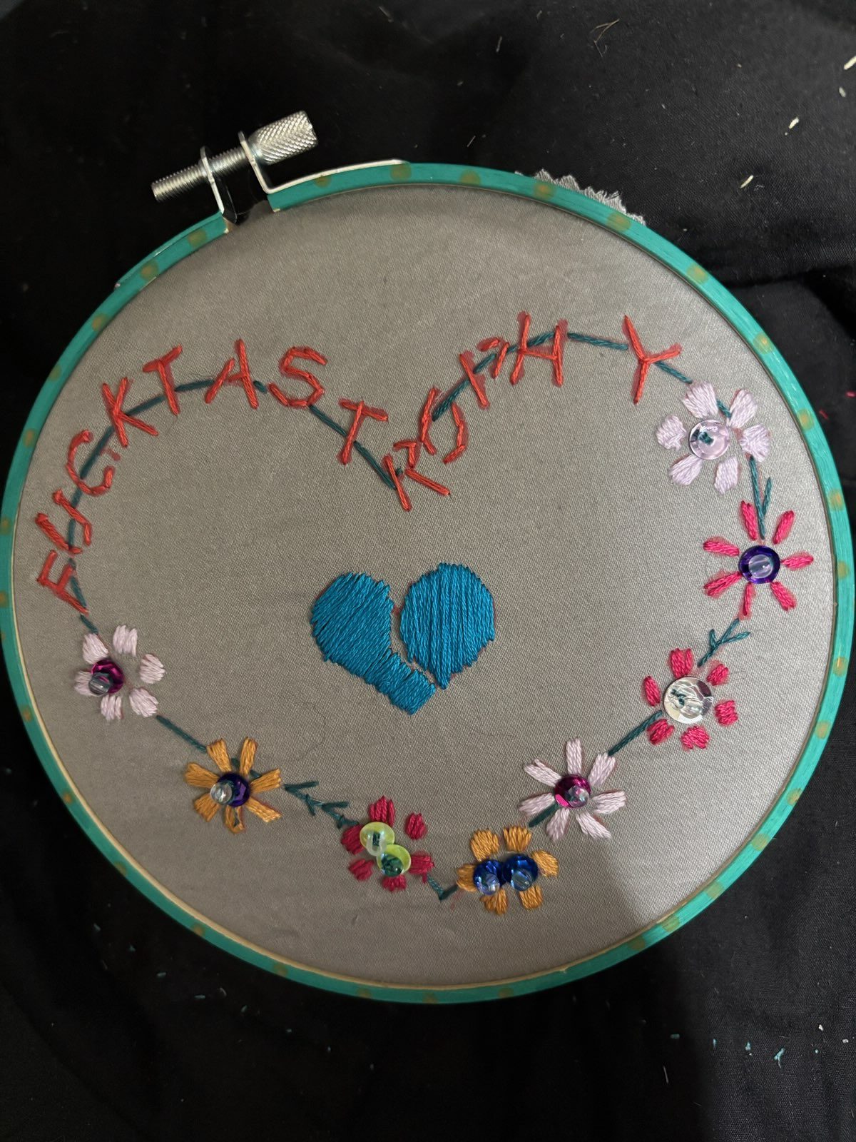 A single Fucktastrophy hoop close up: grey satin in a teal hoop flecked with gold, the word stitched in orange curving into a heart around a teal broken heart, ringed with pink, white and orange flowers with sequin and bead centers.