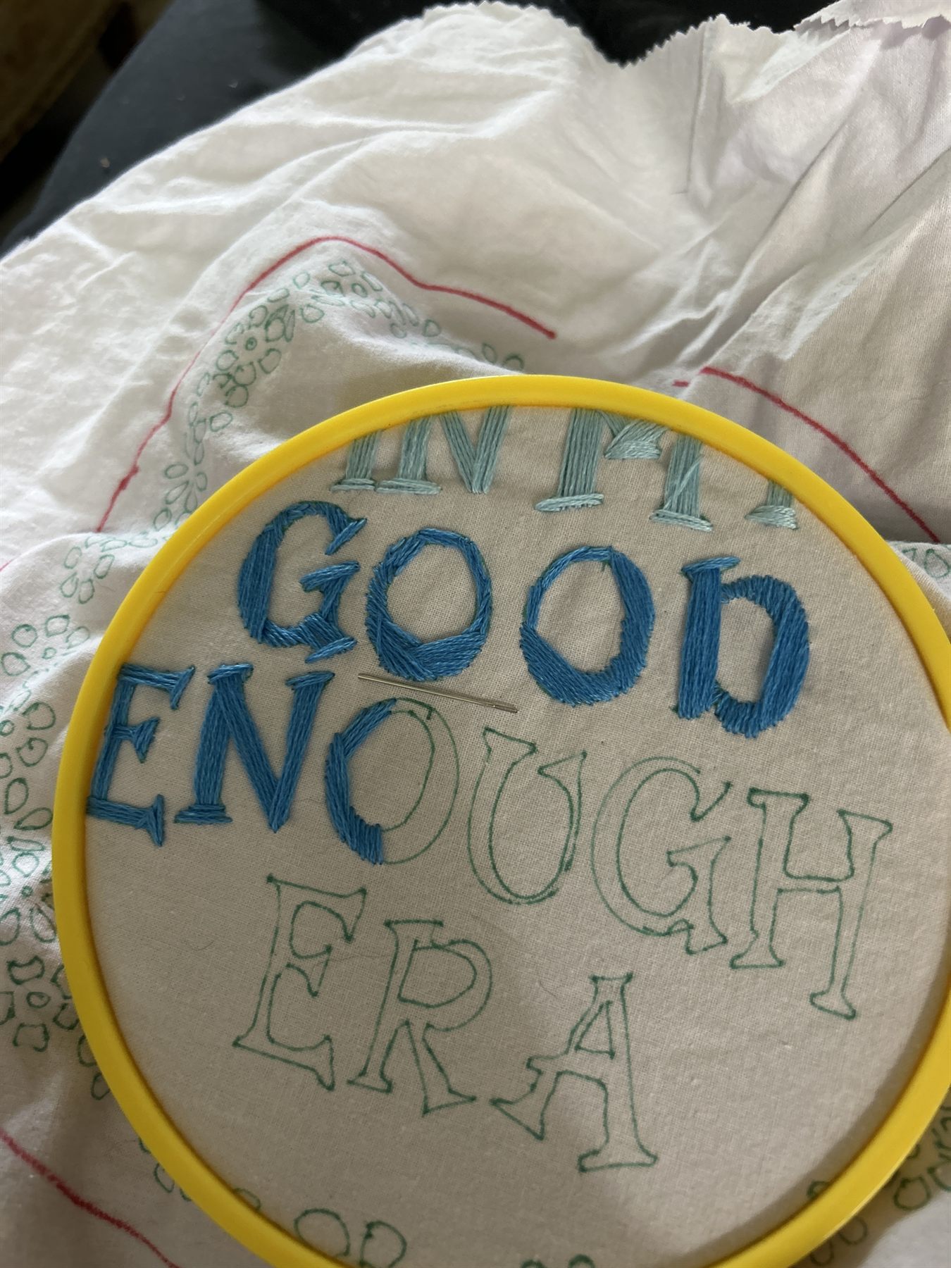 Embroidery hoop with the words In My Good Enough Era, partly stitched in blue floss on calico.
